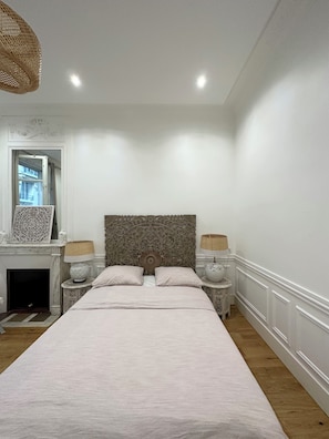 Quarto com cama grande, duas mesas de cabeceira, cabeceira decorativa, lareira e janela com cortinas.