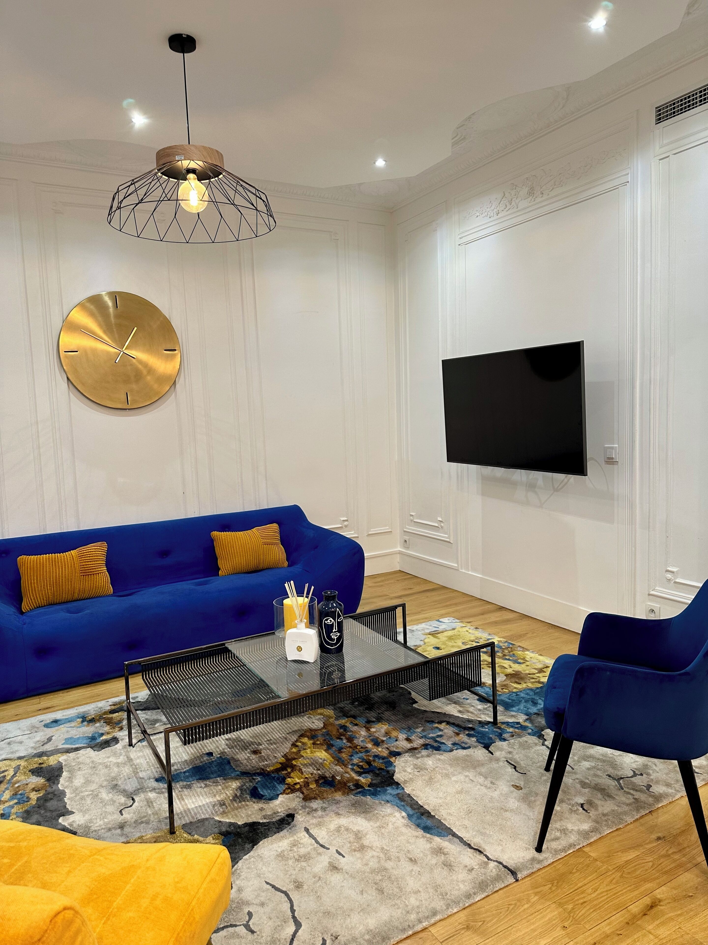 A modern living room with a blue sofa, a round wall clock, a flat-screen TV, and a coffee table with decorative items.