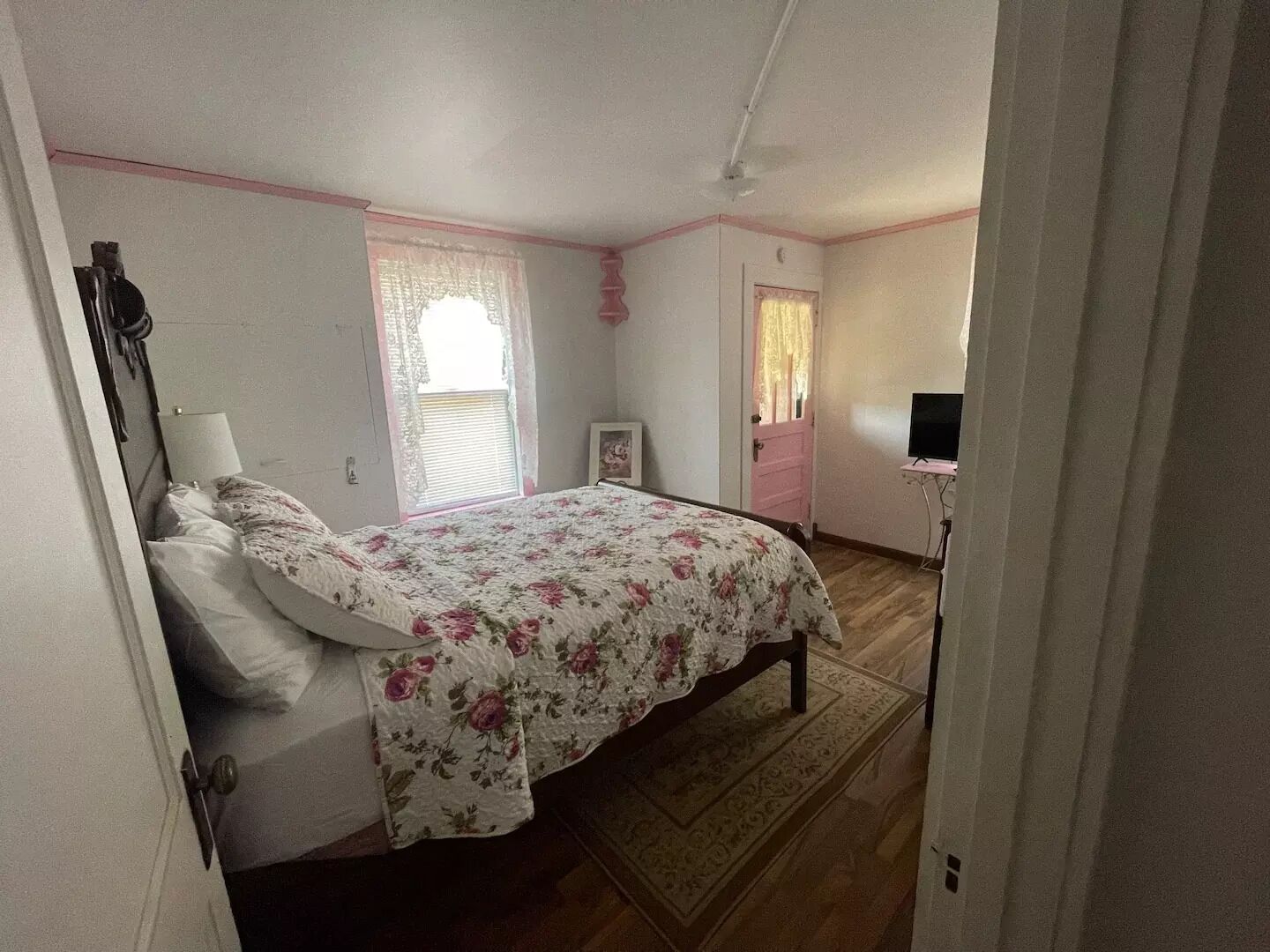 Un dormitorio con cama, ventana con cortinas de encaje, puerta y televisor sobre un mueble.