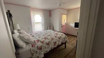 Un dormitorio con cama, ventana con cortinas de encaje, puerta y televisor sobre un mueble.