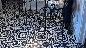 Une pièce avec un sol à motifs, deux chaises dotées d’assises rembourrées, une petite table sur laquelle est posé un vase, et un luminaire fixé au mur.