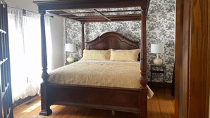 Un lit à baldaquin avec une structure en bois et une tête de lit ornée de motifs floraux. Il y a deux tables de chevet avec des lampes, et la pièce est dotée d’un plancher en bois.