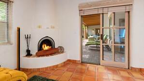A cozy room with a fireplace, a yellow ottoman, and a view through a glass door to an outdoor seating area.