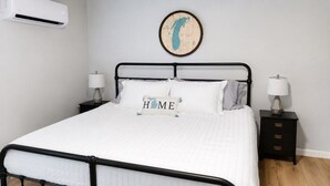 A neatly made bed with a 'HOME' pillow, two bedside tables with lamps, a wall-mounted air conditioner, and a map decoration. - Modern Private 2 Room Suite in Boutique Style Hotel - Room #6 (Muskegon)
