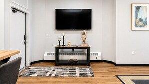 A living room with a flat-screen TV mounted on the wall, a black console table with decorative items, a patterned rug, and a white radiator. - Modern Private 2 Room Suite in Boutique Style Hotel - Room #6 (Muskegon)