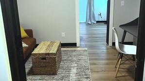 Une pièce avec un canapé marron, un pouf en osier, une chaise blanche et un tapis à motifs.