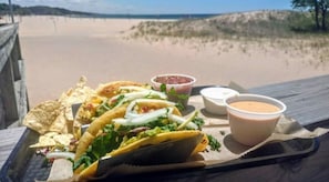A tray with tacos, chips, and two dipping sauces on a wooden deck overlooking a beach.