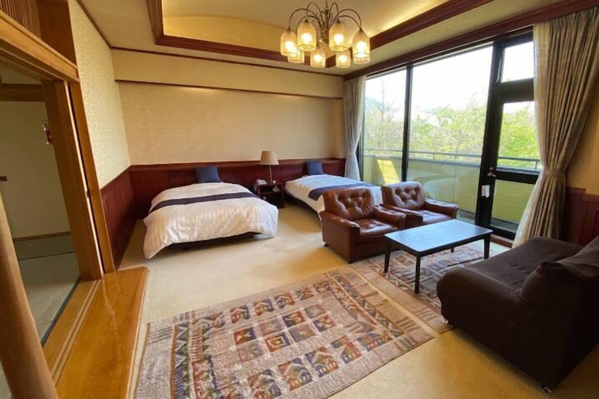 A hotel room with a bed, two armchairs, a sofa, a small table, a patterned rug, and a balcony with curtains.