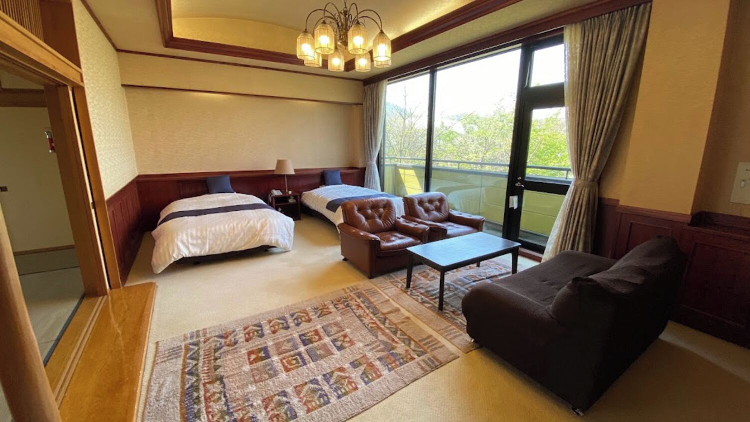 A hotel room with a large bed, a sofa, a chair, a small table, and a patterned rug.