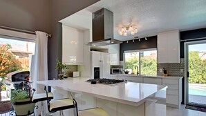 A modern kitchen with a large island, bar stools, and a view of a backyard with a barbecue. - Palm Springs Perfection - Orange Pop Palm Springs (Palm Springs)