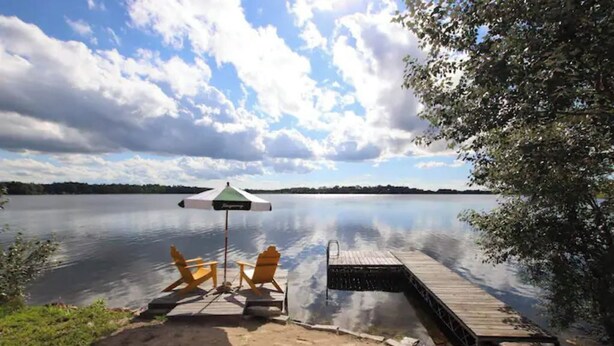 A lakeside view with a dock extending into calm water, two yellow chairs, and a green and white umbrella.