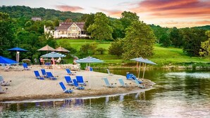A lakeside resort with sun loungers and umbrellas, a house in the background, and a grassy area.