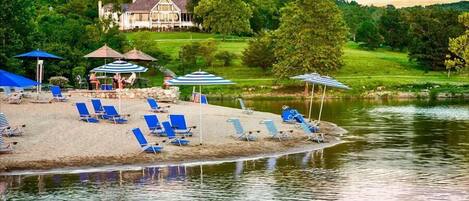 A lakeside resort with sun loungers and umbrellas, a house in the background, and a grassy area.