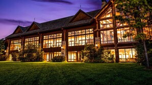 A large, multi-story log cabin with extensive glass windows, surrounded by a well-maintained lawn.