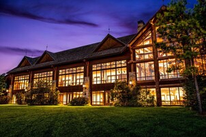 A large, multi-story log cabin with extensive glass windows, surrounded by a well-maintained lawn.