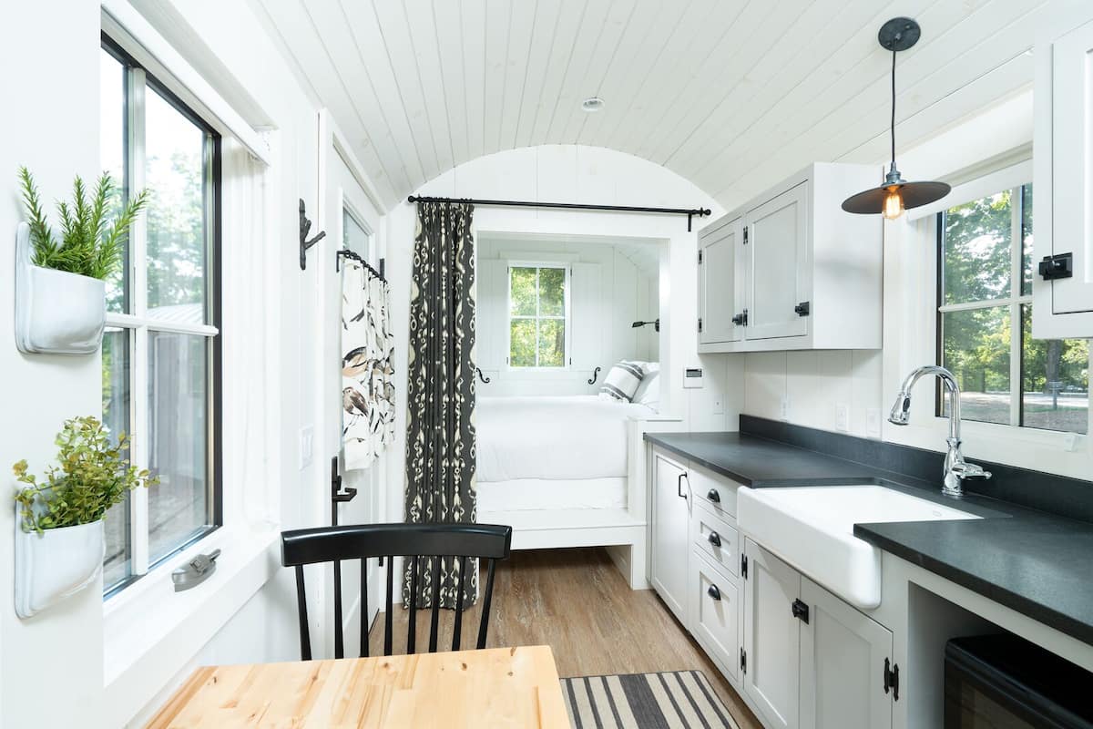 A modern kitchen with white cabinets, a wooden dining table, and a view into a bedroom.