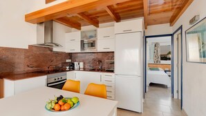 Eine kompakte Küche mit weißen Schränken, einer Mikrowelle und einem Kühlschrank. Ein Esstisch mit Obstschale und orangefarbenen Stühlen steht bereit. Die Küche hat eine Holzbalkendecke und bietet einen Blick in ein Schlafzimmer durch eine offene Tür.