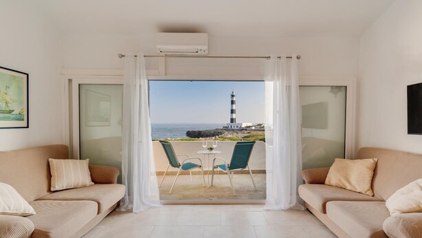 Ein Zimmer mit zwei beigefarbenen Sofas, einer Glastür, die auf einen Balkon mit Blick auf einen Leuchtturm führt, und einem kleinen Tisch mit Stühlen.