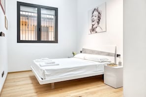 A single bed with a white mattress and pillows, a bedside table with a lamp, a framed picture on the wall, and a window with metal bars.