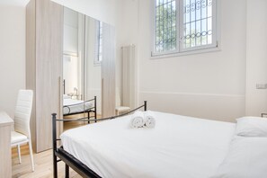 Quarto com cama, poltrona, escrivaninha, radiador e janela com grades.