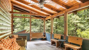 A wooden cabin with a large window overlooking a forest. Inside, there is a wicker sofa with orange cushions, a wooden bench, and a small table with a chair. - Twin Creeks Lodge by Avantstay 33 Acre Retreat (Brevard)