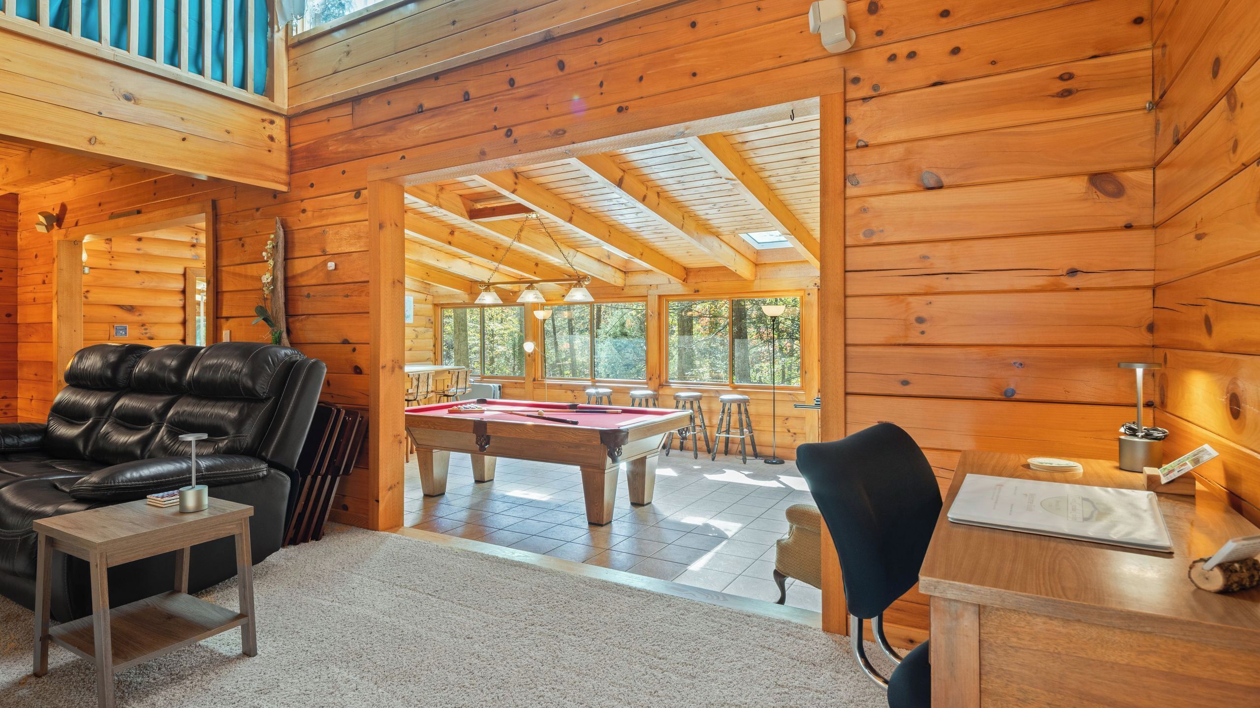 Interior de una cabaña de madera con mesa de billar, sofá de cuero y comedor con luz natural.