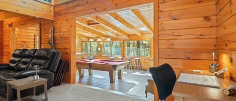 Interior de una cabaña de madera con mesa de billar, sofá de cuero y comedor con luz natural.
