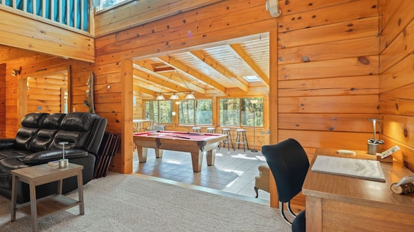 A wooden cabin interior with a pool table, leather sofa, and a dining area with natural light.
