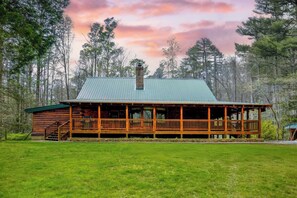 A log cabin with a covered porch and a chimney, surrounded by a grassy field and trees. - Twin Creeks Lodge by Avantstay 33 Acre Retreat (Brevard)