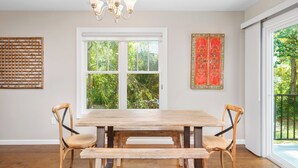 A dining area with a wooden table and chairs, a window with a view of greenery, and a decorative wall piece. - Little Sycamore by Avantstay Premier Location + Patio (Asheville)