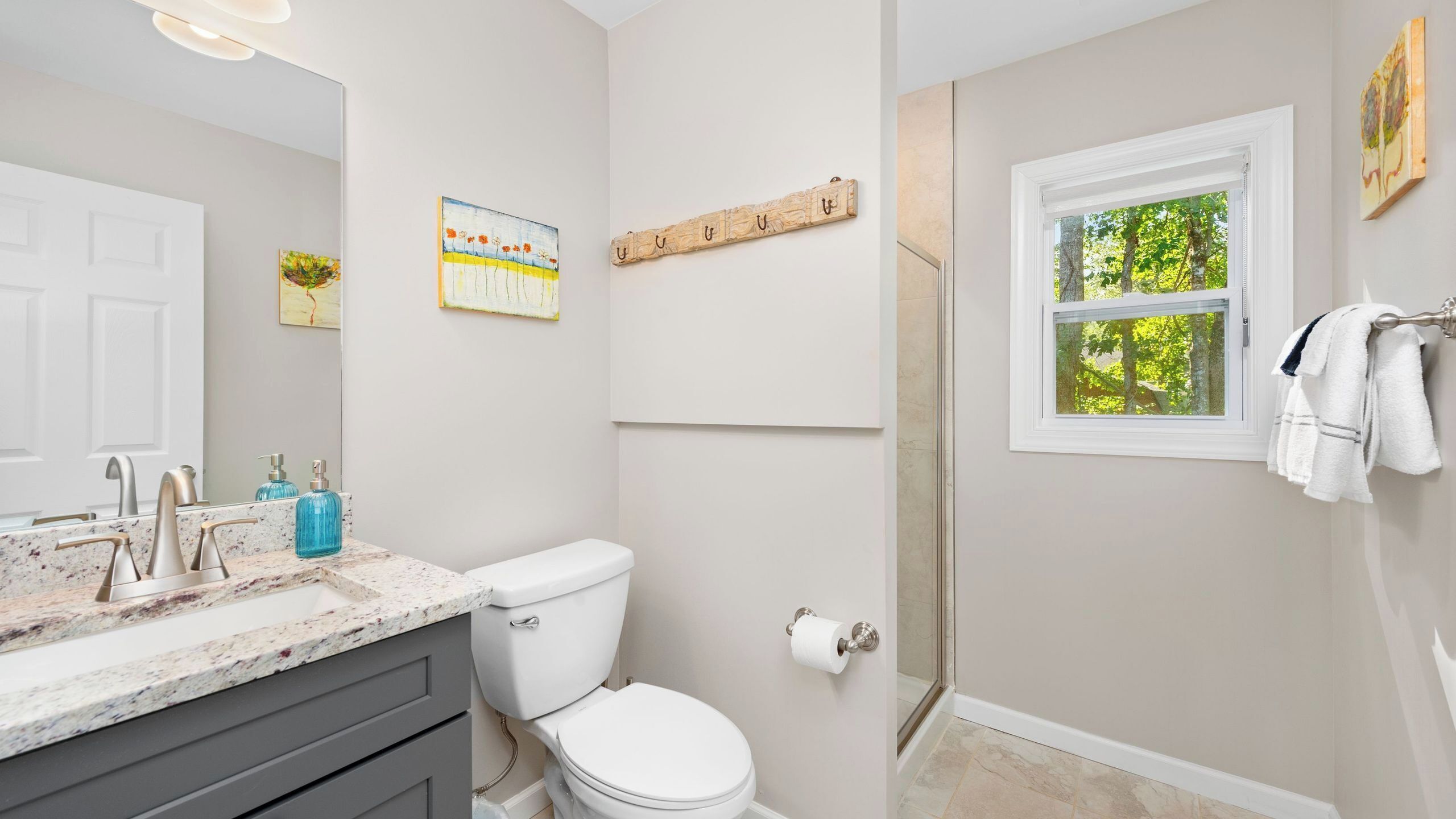 A bathroom with a toilet, sink, and shower. There is a window with a view of trees and a towel rack with towels.