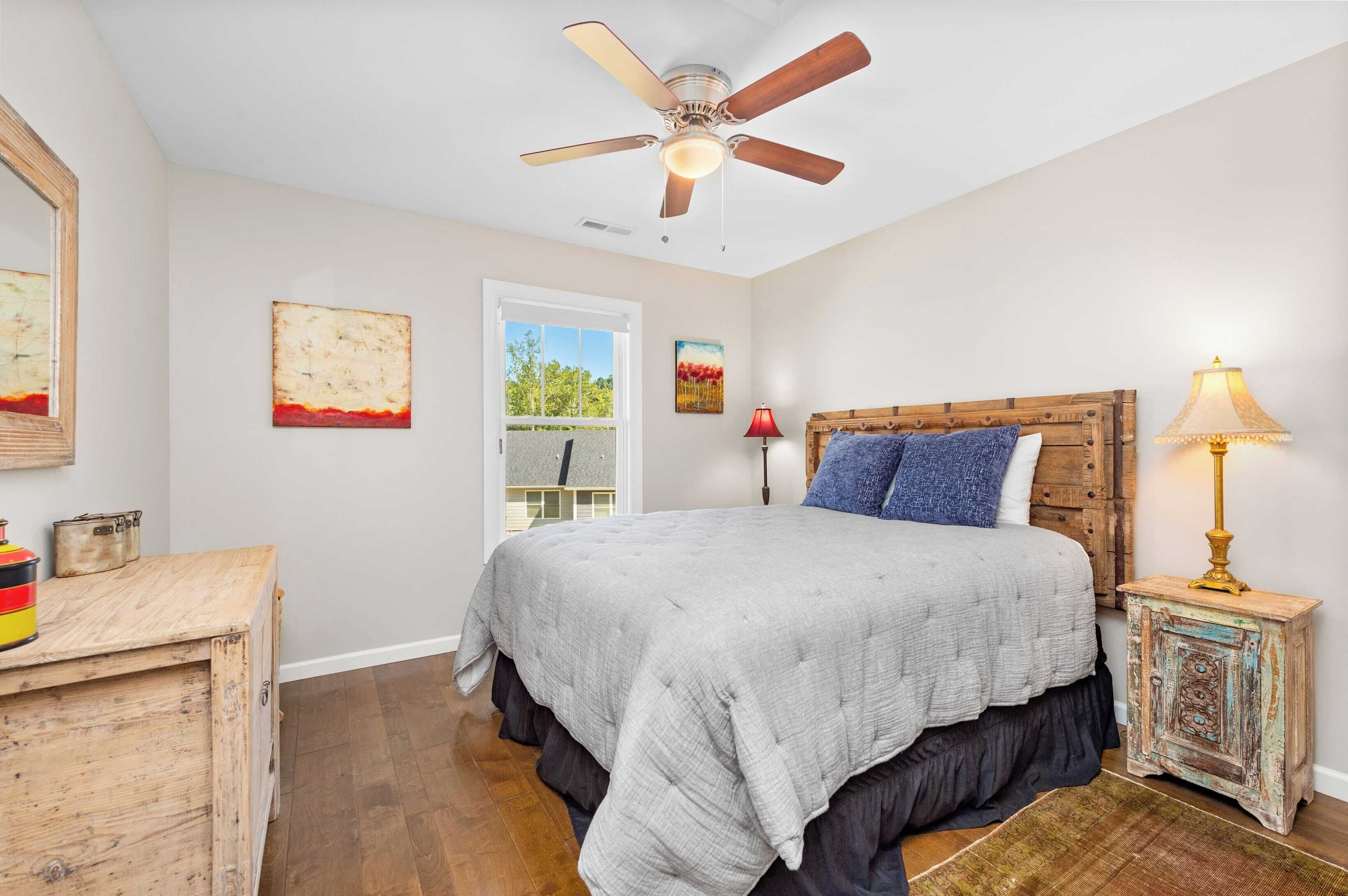 A bedroom with a bed, a ceiling fan, a nightstand with a lamp, and artwork on the walls.