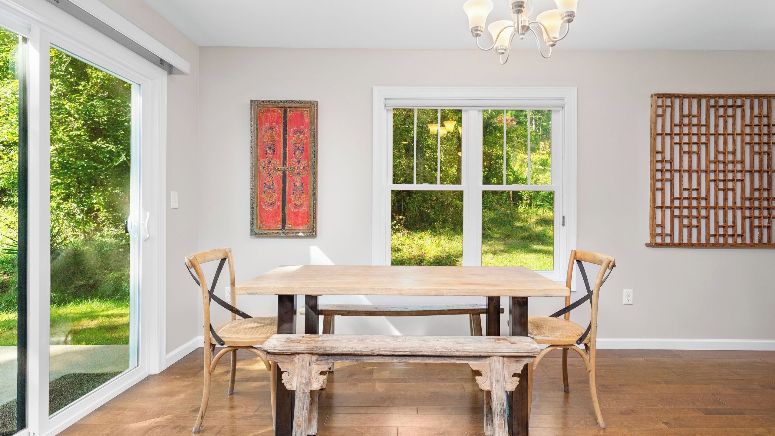A dining area with a wooden table and chairs, a chandelier, and a view of greenery through large windows.