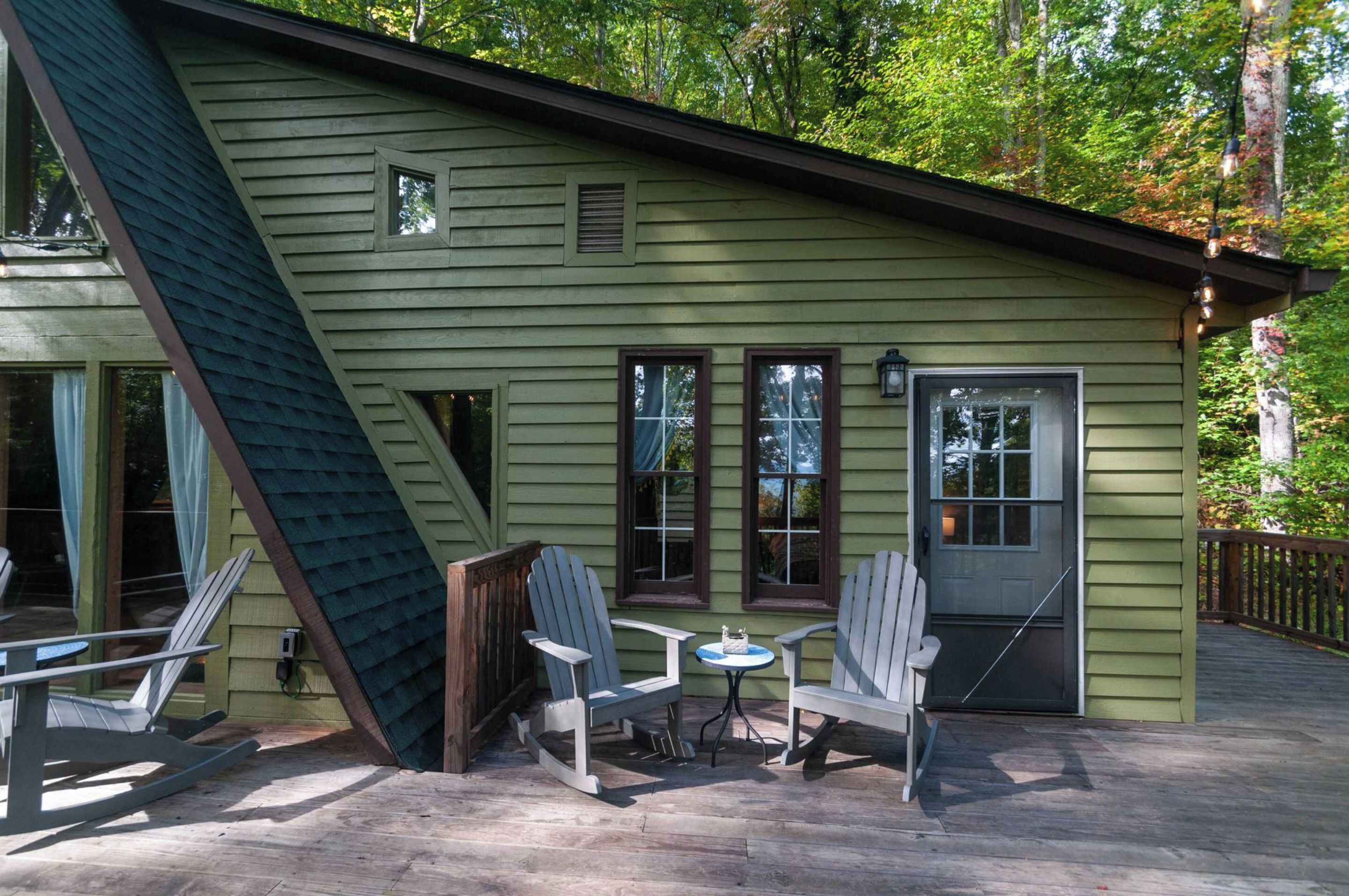 Ein grünes Gebäude mit einer Terrasse, Adirondack-Stühlen, einem kleinen Tisch und einer Tür.