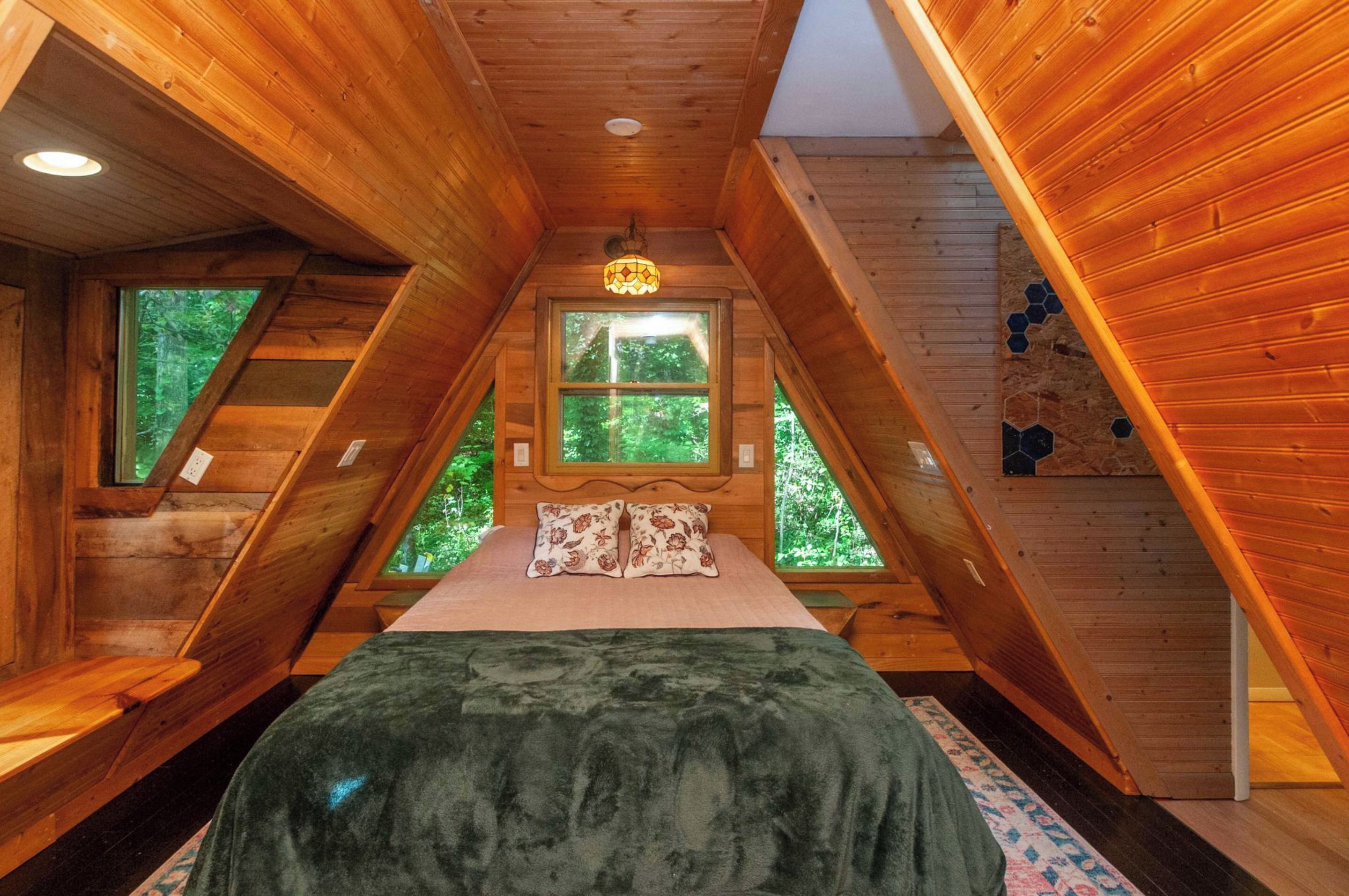 Een houten chalet met een slaapkamer op een entresol, een bed met bloemenkussens, en uitzicht op het bos door grote ramen.
