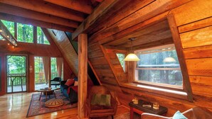 A cozy log cabin interior with a wooden ceiling, a large window with a view of the forest, a comfortable seating area with a sofa and chairs, and a small table with a lamp. - Firepit + Mountain Views A-frame in Fairview Honeycomb Hideaway by Avantstay (Fairview)