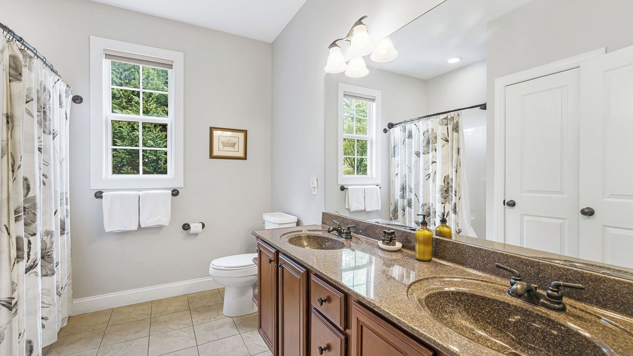 Une salle de bain avec deux lavabos, des toilettes et une douche. Il y a des fenêtres avec des rideaux, un tableau encadré au mur et un miroir au-dessus des lavabos.