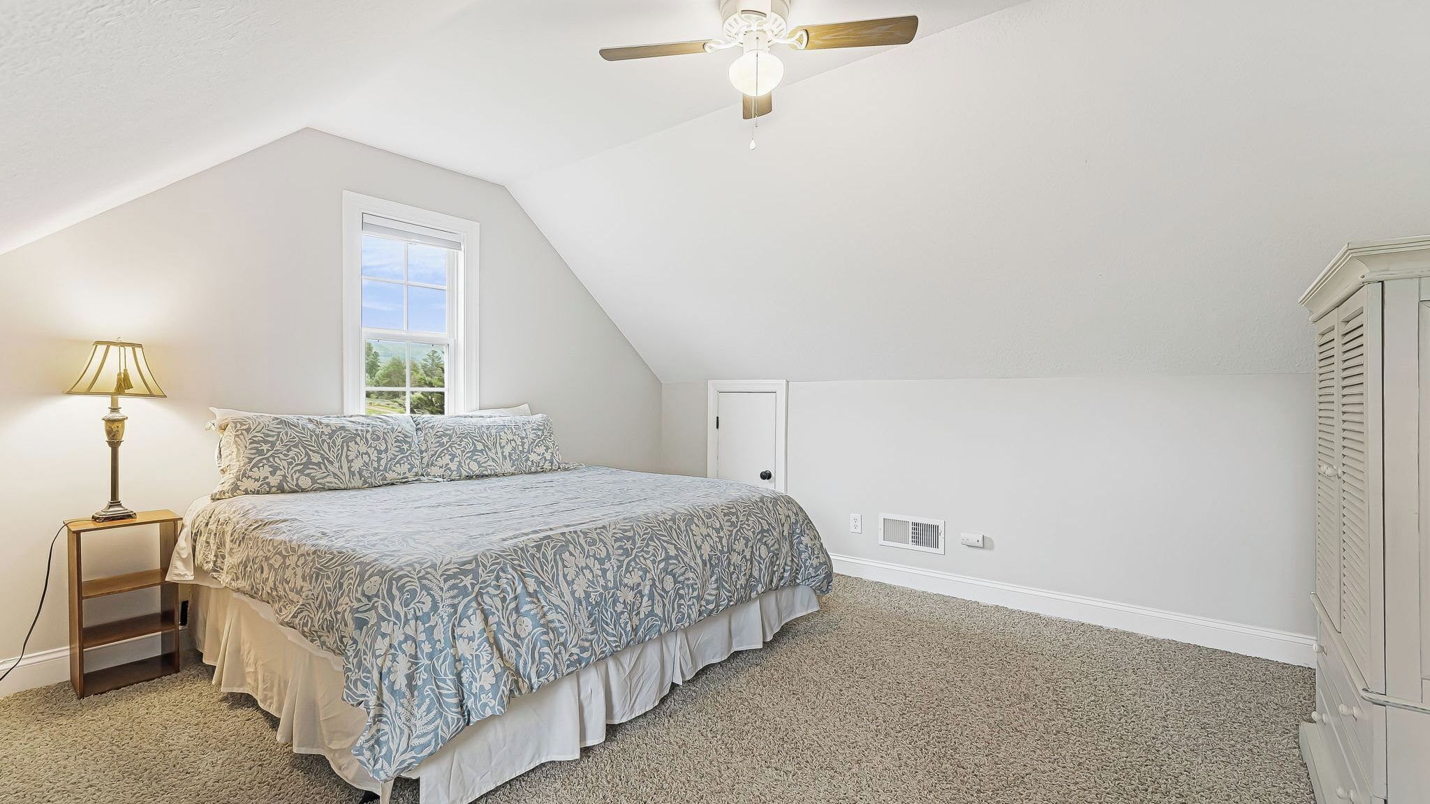 A bedroom with a sloped ceiling, a bed with floral bedding, a nightstand with a lamp, a window, and a door.