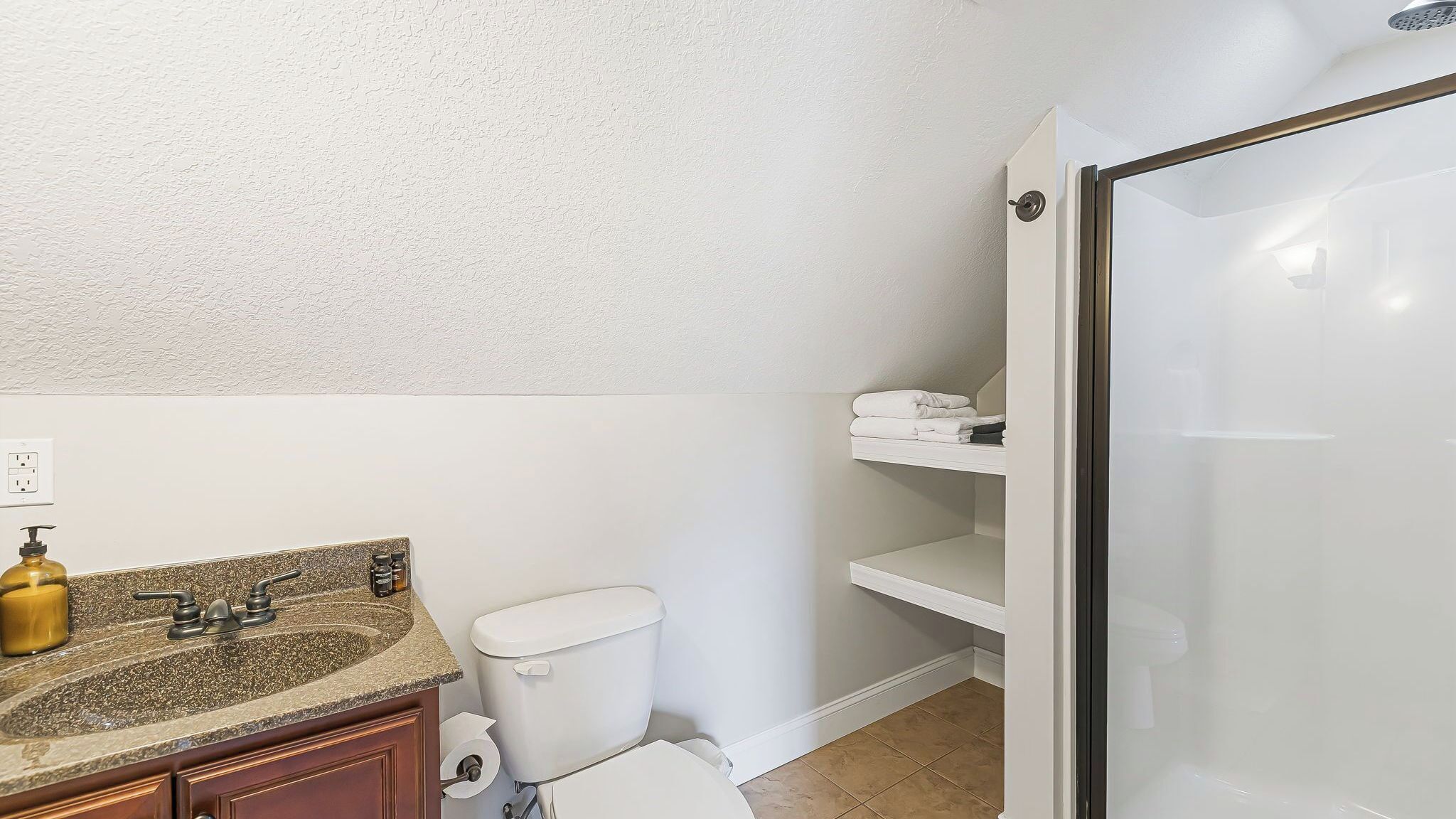 A bathroom with a granite countertop, a toilet, a shower, and a shelf with towels.