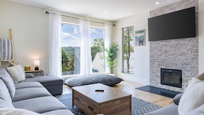 A modern living room with a grey sofa, a wooden coffee table, a fireplace, and large windows with a view of trees. - Hot Tub, Fire Pit, Mountain Views, 10min to AVL Modern Escape by Avanstay (Asheville)