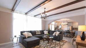 Amplia sala de estar con un sofá modular grande, una mesa de centro de vidrio y un tapete estampado. Hay una araña y se ve la cocina a través de un arco.