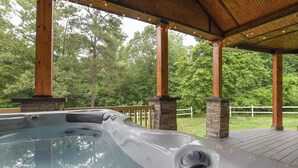 Un jacuzzi bajo una terraza de madera, con pilares de piedra, rodeado por una valla blanca y árboles.