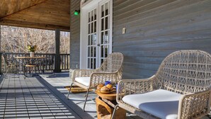 A covered porch with wicker chairs and a table, a wooden deck, and a house with a window and door. - Hamersmith Hollow Retreat by Avantstay Mountain Views (Mars Hill)