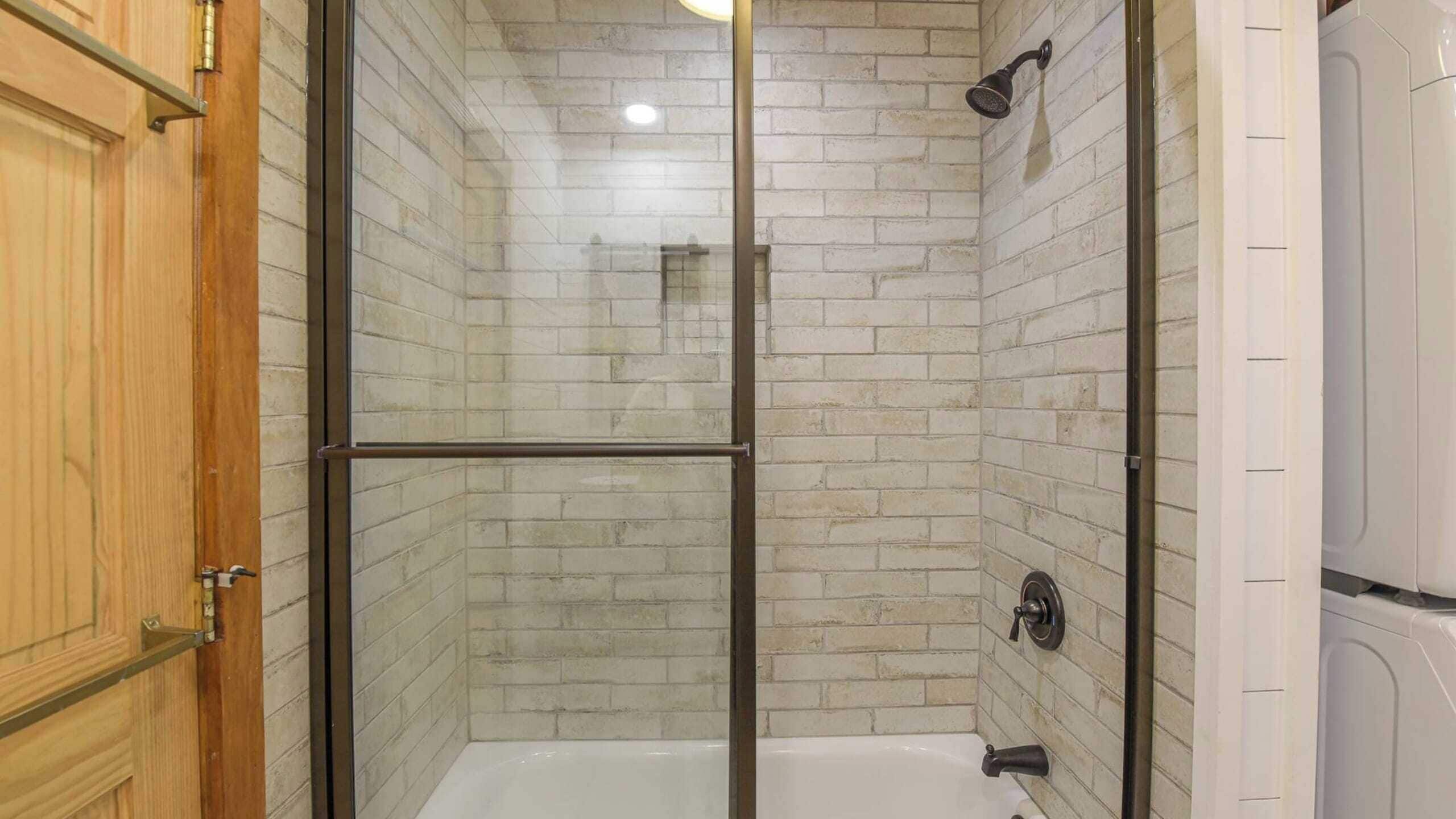 A glass-enclosed shower stall with a white bathtub, tiled walls, and a wooden door.