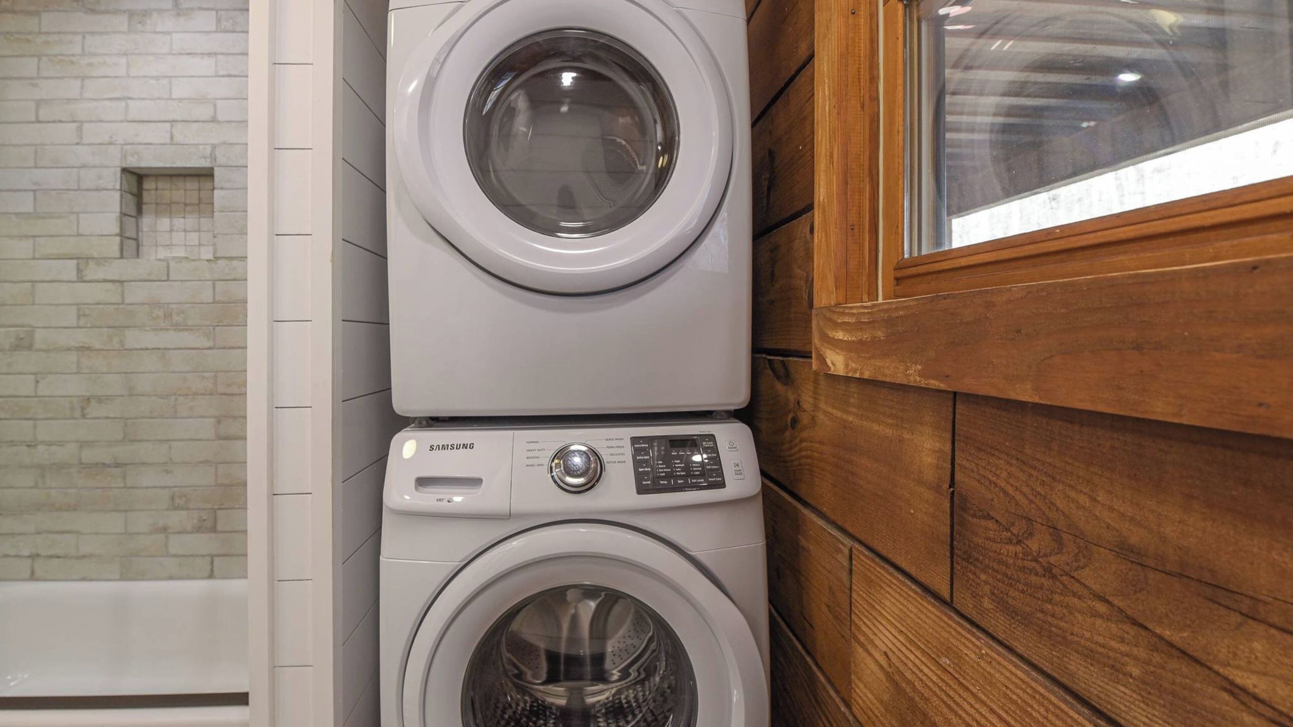 Een gestapelde wasmachine en droger in een badkamer met een bakstenen muur en houten lambrisering.