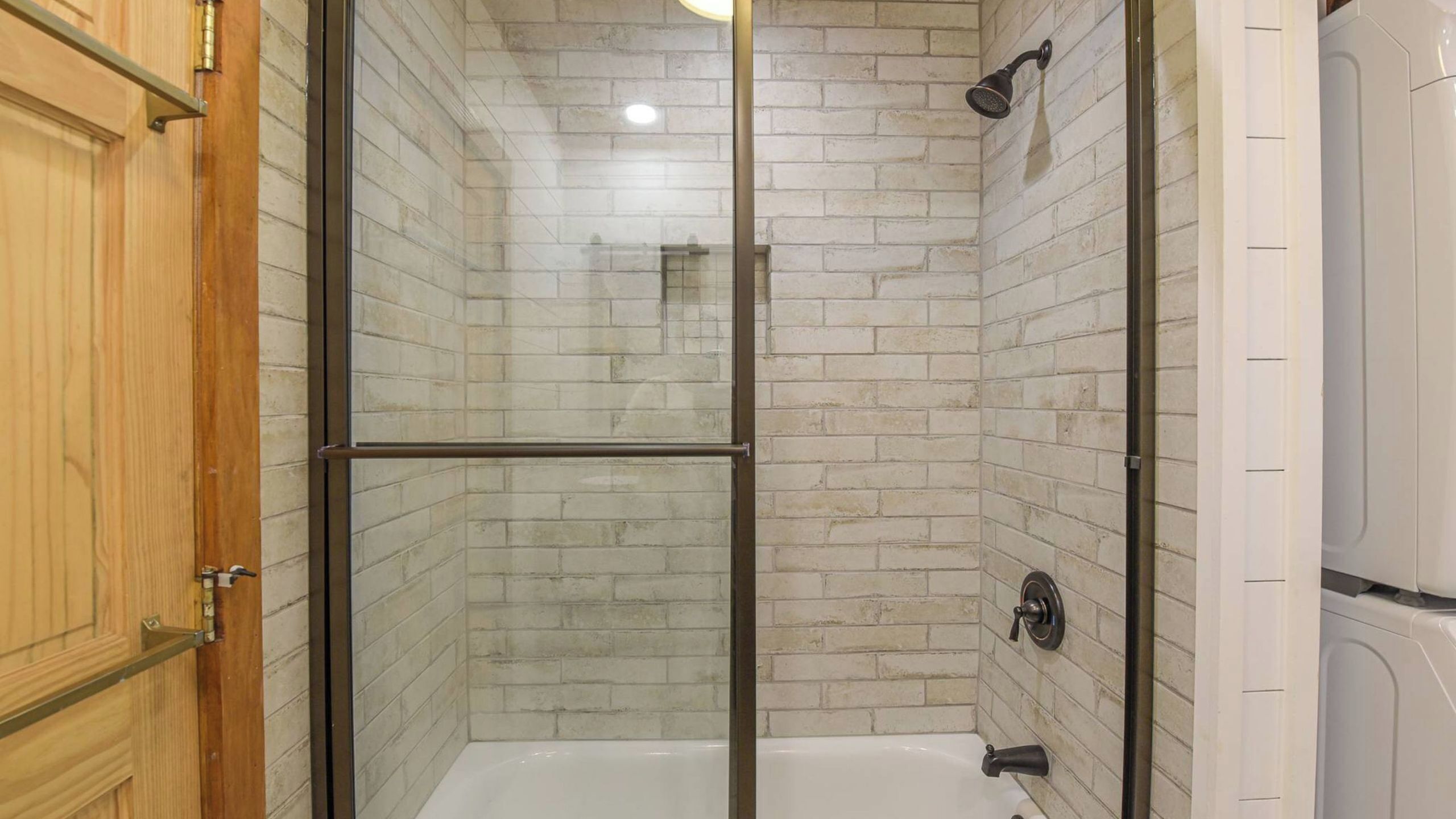 A glass-enclosed shower stall with a white bathtub, wooden door, and a showerhead.