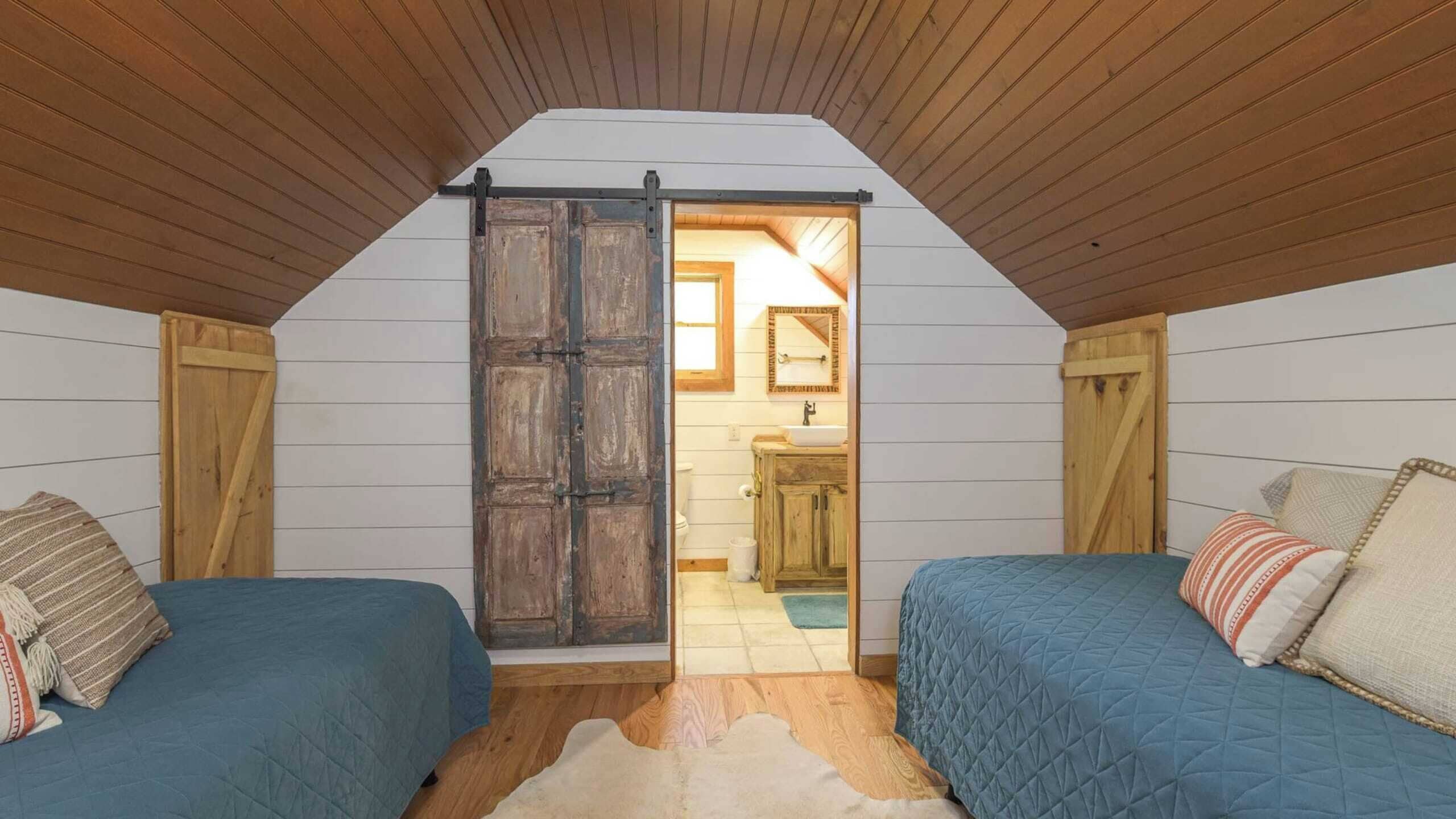 Un dormitorio acogedor en el ático, con dos camas, paredes de madera y una puerta rústica que da al baño.
