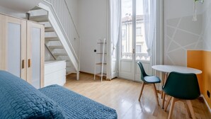Um quarto com cama, escada, janela com cortinas, uma pequena mesa com cadeiras e uma planta.