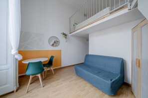 Une pièce de taille modeste, comprenant un canapé, une petite table avec des chaises, un miroir et une plante suspendue.