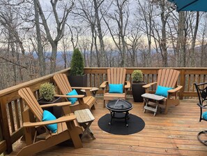 A wooden deck with Adirondack chairs and a round table, surrounded by potted plants and overlooking a forested area.